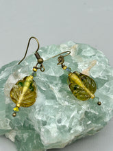 Load image into Gallery viewer, This pair of earrings features a dark amber and green tone, pressed glass leaf charm in an iridescent finish that gives off subtle hues of gold. Complemented with light ming green glass beaded earrings, this set is sure to sparkle.   Measurements: 1 1/4" length