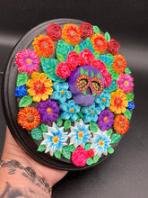 Load image into Gallery viewer, Frida Kahlo inspired calavera with a black wood frame, this Day of the Dead wall decor piece is perfect for adding a traditional Mexican folk art touch to any home. Add a unique touch of style to your home with this intricately detailed, Frida Kahlo-inspired calavera clay wall art. The design features an eye-catching crown of flowers, plus a vibrant bouquet, perfect for any Day of the Dead celebration. Bring a bright and colorful piece of art to your home with this stunning wall hanging.