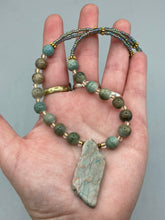 Load image into Gallery viewer, This exquisite necklace showcases earthy tones of mossy green ceramic beads, complemented by a gray toned howlite stone chip pendant. To add a sparkle, I interspersed gold-tone glass beads between each ceramic one. Two delicate strands feature small glass beads with hues of blue, gold, and purple. These are bridged by flower-shaped gold plated beads, which then unite with the round ceramic beads. This alluring earth-tone combination makes a stunning accessory. Necklace is 17”