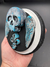 Load image into Gallery viewer, This handcrafted Dia De Los Muertos Calavera wall art decor is a unique way to bring life to your home and celebrates Mexican culture. Made with polymere clay and painted on a wooden frame, the beautiful blue skull is finished with wings and fleur de lis embellishments for a sophisticated look. Perfect for any room! Measurements: D 4"