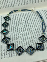 Load image into Gallery viewer, This two-strand necklace, crafted from aqua-blue and silver toned glass beads, is a stunning statement piece. The addition of faceted triangle-shaped glass beads tucked between hematite square beads adds an exquisite and unique flair. The necklace gracefully drapes around the neck, bringing an air of bold and elegant flair to any ensemble it complements. 15” in length, can be extended up to 17"