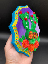 Load image into Gallery viewer, This handcrafted polymer clay wall art features a wood frame painted in a gradient from light to dark purple, creating a stunning sunset look. With a bright green skull cactus plant and bright red blooming flowers, this piece is sure to make a statement. Measurements: H 7" x W 5” x D 1 1/2"