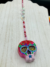Load image into Gallery viewer, Nature inspired hand crafted polymer clay skull is painted in ombre colors from red, to light pink and decorated with flowers on back and over eyes. Three hearts rainbow hematite bead on forehead. Light pink glass beaded chain with iridescent crystals. A perfect hanging Day of the Dead art piece to add to your ofrenda, hang next to your display of crystals or witchy altar, add to your skull collection or hang as an art piece on the wall or near a window. Measurements: H 1 1/2” x W 1 1/2” x D 3/4”