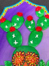 Load image into Gallery viewer, This handcrafted polymer clay wall art features a wood frame painted in a gradient from light to dark purple, creating a stunning sunset look. With a bright green skull cactus plant and bright red blooming flowers, this piece is sure to make a statement. Measurements: H 7" x W 5” x D 1 1/2"