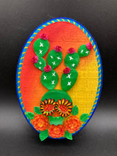 Load image into Gallery viewer, This handcrafted polymer clay wall art features a wood frame painted in a gradient from light yellow to dark red, creating a stunning sunset look. With a bright green skull cactus plant and bright pink blooming flowers. Boasting vivid marigold blooms and a deep royal blue frame to make a bold contrast with the warm background hue. this piece is sure to make a statement. Measurements: H 7" x W 5” x D 1 1/2"