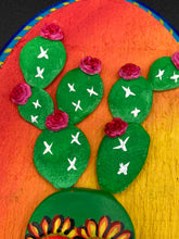 Load image into Gallery viewer, This handcrafted polymer clay wall art features a wood frame painted in a gradient from light yellow to dark red, creating a stunning sunset look. With a bright green skull cactus plant and bright pink blooming flowers. Boasting vivid marigold blooms and a deep royal blue frame to make a bold contrast with the warm background hue. this piece is sure to make a statement. Measurements: H 7" x W 5” x D 1 1/2"