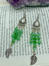 Load image into Gallery viewer, This large set of chandelier earrings features three long strands of shimmering glass beads in various light green and blue hues, accented with an intricate silver-plated leaf charm at the bottom for an added touch of movement. Versatile and perfect for whimsical, hippie-inspired, nature-loving looks. Measurements: 3 3/4” ” in length