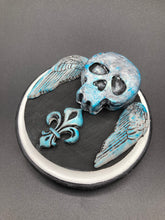 Load image into Gallery viewer, This handcrafted Dia De Los Muertos Calavera wall art decor is a unique way to bring life to your home and celebrates Mexican culture. Made with polymere clay and painted on a wooden frame, the beautiful blue skull is finished with wings and fleur de lis embellishments for a sophisticated look. Perfect for any room! Measurements: D 4"