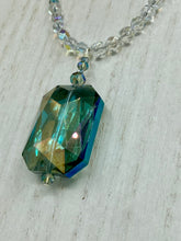 Load image into Gallery viewer, This stunning aqua blue and silver glass beaded necklace is a bold statement piece, composed of an adjustable necklace featuring blues, silver, and iridescent beads. Its shorter, collar style strand contains two-hole iridescent beads to keep its shape and adds a generous helping of iridescent teardrop glass beads for maximum sparkle. Its accompanying large, rectangular faceted pendant has a shimmering mix of gold, green, pink, and blue hues, and elegantly drapes across the neck.