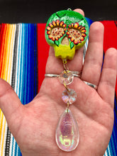 Load image into Gallery viewer, Nature inspired hand crafted polymer clay skull is painted in ombre colors from dark green to light yellow and decorated with flowers on back and over eyes. Light gold glass beaded chain with iridescent crystals. A perfect hanging Day of the Dead art piece to add to your ofrenda, hang next to your display of crystals or witchy altar, add to your skull collection or hang as an art piece on the wall or near a window. Measurements: H 1 1/2” x W 1 1/2” x D 3/4”