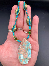 Load image into Gallery viewer, This stunning two-strand necklace features an array of glass beads in hues of green and blue turquoise, gold and green for a vibrant, colorful and bold look. The centerpiece is a large howlite stone chip dyed in turquoise color, accented by the bronze patina cowry beads. Measurements: Necklace is 17”