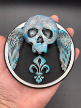 Load image into Gallery viewer, This handcrafted Dia De Los Muertos Calavera wall art decor is a unique way to bring life to your home and celebrates Mexican culture. Made with polymere clay and painted on a wooden frame, the beautiful blue skull is finished with wings and fleur de lis embellishments for a sophisticated look. Perfect for any room! Measurements: D 4"