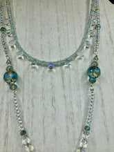 Load image into Gallery viewer, This stunning aqua blue and silver glass beaded necklace is a bold statement piece, composed of an adjustable necklace featuring blues, silver, and iridescent beads. Its shorter, collar style strand contains two-hole iridescent beads to keep its shape and adds a generous helping of iridescent teardrop glass beads for maximum sparkle. Its accompanying large, rectangular faceted pendant has a shimmering mix of gold, green, pink, and blue hues, and elegantly drapes across the neck.