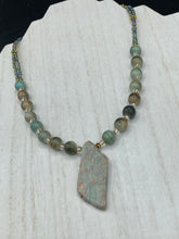 Load image into Gallery viewer, This exquisite necklace showcases earthy tones of mossy green ceramic beads, complemented by a gray toned howlite stone chip pendant. To add a sparkle, I interspersed gold-tone glass beads between each ceramic one. Two delicate strands feature small glass beads with hues of blue, gold, and purple. These are bridged by flower-shaped gold plated beads, which then unite with the round ceramic beads. This alluring earth-tone combination makes a stunning accessory. Necklace is 17”