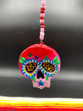 Load image into Gallery viewer, Nature inspired hand crafted polymer clay skull is painted in ombre colors from red, to light pink and decorated with flowers on back and over eyes. Three hearts rainbow hematite bead on forehead. Light pink glass beaded chain with iridescent crystals. A perfect hanging Day of the Dead art piece to add to your ofrenda, hang next to your display of crystals or witchy altar, add to your skull collection or hang as an art piece on the wall or near a window. Measurements: H 1 1/2” x W 1 1/2” x D 3/4”
