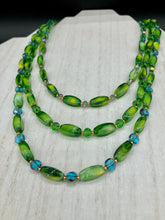 Load image into Gallery viewer, This adjustable glass beaded necklace features a combination of vivid green, blue, and gold hues and a mixture of shapes, creating an eye-catching spectrum of colors and a complex look. The rectangular green glass beads form the predominant part of the three-strand piece, which offers a bold and elegant fashion statement that is ideal for the Spring season. Measurements: Necklace is 17” in length, can be adjusted up to 19" in length