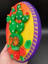 Load image into Gallery viewer, This handcrafted polymer clay wall art features a wood frame painted in a gradient from light yellow to dark red, creating a stunning sunset look. With a bright green skull cactus plant and bright yellow/red blooming flowers. Boasting vivid marigold blooms and a deep purple frame to make a bold contrast with the warm background hue. this piece is sure to make a statement. Measurements: H 7" x W 5” x D 1 1/2"