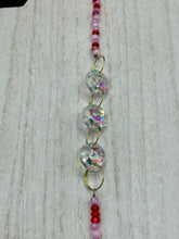 Load image into Gallery viewer, Nature inspired hand crafted polymer clay skull is painted in ombre colors from red, to light pink and decorated with flowers on back and over eyes. Three hearts rainbow hematite bead on forehead. Light pink glass beaded chain with iridescent crystals. A perfect hanging Day of the Dead art piece to add to your ofrenda, hang next to your display of crystals or witchy altar, add to your skull collection or hang as an art piece on the wall or near a window. Measurements: H 1 1/2” x W 1 1/2” x D 3/4”