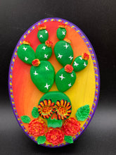 Load image into Gallery viewer, This handcrafted polymer clay wall art features a wood frame painted in a gradient from light yellow to dark red, creating a stunning sunset look. With a bright green skull cactus plant and bright yellow/red blooming flowers. Boasting vivid marigold blooms and a deep purple frame to make a bold contrast with the warm background hue. this piece is sure to make a statement. Measurements: H 7" x W 5” x D 1 1/2"
