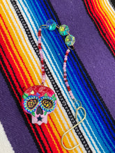 Load image into Gallery viewer, Nature inspired hand crafted polymer clay skull is painted in ombre colors from red, to light pink and decorated with flowers on back and over eyes. Three hearts rainbow hematite bead on forehead. Light pink glass beaded chain with iridescent crystals. A perfect hanging Day of the Dead art piece to add to your ofrenda, hang next to your display of crystals or witchy altar, add to your skull collection or hang as an art piece on the wall or near a window. Measurements: H 1 1/2” x W 1 1/2” x D 3/4”