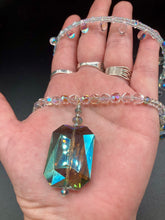Load image into Gallery viewer, This stunning aqua blue and silver glass beaded necklace is a bold statement piece, composed of an adjustable necklace featuring blues, silver, and iridescent beads. Its shorter, collar style strand contains two-hole iridescent beads to keep its shape and adds a generous helping of iridescent teardrop glass beads for maximum sparkle. Its accompanying large, rectangular faceted pendant has a shimmering mix of gold, green, pink, and blue hues, and elegantly drapes across the neck.