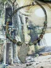 Load image into Gallery viewer, This exquisite necklace showcases earthy tones of mossy green ceramic beads, complemented by a gray toned howlite stone chip pendant. To add a sparkle, I interspersed gold-tone glass beads between each ceramic one. Two delicate strands feature small glass beads with hues of blue, gold, and purple. These are bridged by flower-shaped gold plated beads, which then unite with the round ceramic beads. This alluring earth-tone combination makes a stunning accessory. Necklace is 17”