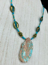 Load image into Gallery viewer, This stunning two-strand necklace features an array of glass beads in hues of green and blue turquoise, gold and green for a vibrant, colorful and bold look. The centerpiece is a large howlite stone chip dyed in turquoise color, accented by the bronze patina cowry beads. Measurements: Necklace is 17”