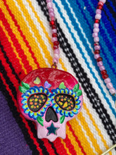 Load image into Gallery viewer, Nature inspired hand crafted polymer clay skull is painted in ombre colors from red, to light pink and decorated with flowers on back and over eyes. Three hearts rainbow hematite bead on forehead. Light pink glass beaded chain with iridescent crystals. A perfect hanging Day of the Dead art piece to add to your ofrenda, hang next to your display of crystals or witchy altar, add to your skull collection or hang as an art piece on the wall or near a window. Measurements: H 1 1/2” x W 1 1/2” x D 3/4”