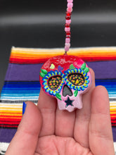 Load image into Gallery viewer, Nature inspired hand crafted polymer clay skull is painted in ombre colors from red, to light pink and decorated with flowers on back and over eyes. Three hearts rainbow hematite bead on forehead. Light pink glass beaded chain with iridescent crystals. A perfect hanging Day of the Dead art piece to add to your ofrenda, hang next to your display of crystals or witchy altar, add to your skull collection or hang as an art piece on the wall or near a window. Measurements: H 1 1/2” x W 1 1/2” x D 3/4”