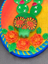 Load image into Gallery viewer, This handcrafted polymer clay wall art features a wood frame painted in a gradient from light yellow to dark red, creating a stunning sunset look. With a bright green skull cactus plant and bright pink blooming flowers. Boasting vivid marigold blooms and a deep royal blue frame to make a bold contrast with the warm background hue. this piece is sure to make a statement. Measurements: H 7" x W 5” x D 1 1/2"