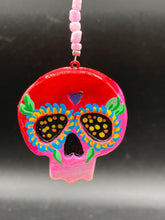 Load image into Gallery viewer, Nature inspired hand crafted polymer clay skull is painted in ombre colors from red, to light pink and decorated with flowers on back and over eyes. Heart rainbow hematite bead on forehead. Light pink glass beaded chain with iridescent crystals. A perfect hanging Day of the Dead art piece to add to your ofrenda, hang next to your display of crystals or witchy altar, add to your skull collection or hang as an art piece on the wall or near a window. Measurements: H 1 1/2” x W 1 1/2” x D 3/4”