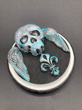 Load image into Gallery viewer, This handcrafted Dia De Los Muertos Calavera wall art decor is a unique way to bring life to your home and celebrates Mexican culture. Made with polymere clay and painted on a wooden frame, the beautiful blue skull is finished with wings and fleur de lis embellishments for a sophisticated look. Perfect for any room! Measurements: D 4"