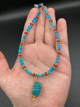 Load image into Gallery viewer, This adjustable choker style necklace features an eye-catching teal wood scarab beetle pendant and glass beaded strands. This dramatic necklace is kept together by double-hole flat matte lava-red beads, providing a collar shape. The necklace artfully wraps around the neck, conveying an elegant and bold style.  Measurements:    Necklace is 14 1/2” in length, can be adjusted up to 16"