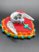 Load image into Gallery viewer, This Memento Mori Wall Art Decor is a unique statement piece to add a pop of color to your walls. Handcrafted with black and white polymer clay, acrylic paint, and a wood frame, the skull artwork is decorated with small turquoise and yellow-orange flowers and a turquoise and gold cross. Perfect for a Day of the Dead altar or a witchy home decor display.
