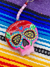 Load image into Gallery viewer, Nature inspired hand crafted polymer clay skull is painted in ombre colors from red, to light pink and decorated with flowers on back and over eyes. Heart rainbow hematite bead on forehead. Light pink glass beaded chain with iridescent crystals. A perfect hanging Day of the Dead art piece to add to your ofrenda, hang next to your display of crystals or witchy altar, add to your skull collection or hang as an art piece on the wall or near a window. Measurements: H 1 1/2” x W 1 1/2” x D 3/4”
