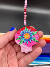 Load image into Gallery viewer, Nature inspired hand crafted polymer clay skull is painted in ombre colors from red, to light pink and decorated with flowers on back and over eyes. Three hearts rainbow hematite bead on forehead. Light pink glass beaded chain with iridescent crystals. A perfect hanging Day of the Dead art piece to add to your ofrenda, hang next to your display of crystals or witchy altar, add to your skull collection or hang as an art piece on the wall or near a window. Measurements: H 1 1/2” x W 1 1/2” x D 3/4”