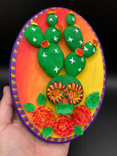 Load image into Gallery viewer, This handcrafted polymer clay wall art features a wood frame painted in a gradient from light yellow to dark red, creating a stunning sunset look. With a bright green skull cactus plant and bright yellow/red blooming flowers. Boasting vivid marigold blooms and a deep purple frame to make a bold contrast with the warm background hue. this piece is sure to make a statement. Measurements: H 7" x W 5” x D 1 1/2"