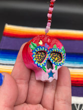 Load image into Gallery viewer, Nature inspired hand crafted polymer clay skull is painted in ombre colors from red, to light pink and decorated with flowers on back and over eyes. Three hearts rainbow hematite bead on forehead. Light pink glass beaded chain with iridescent crystals. A perfect hanging Day of the Dead art piece to add to your ofrenda, hang next to your display of crystals or witchy altar, add to your skull collection or hang as an art piece on the wall or near a window. Measurements: H 1 1/2” x W 1 1/2” x D 3/4”