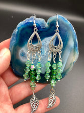 Load image into Gallery viewer, This large set of chandelier earrings features three long strands of shimmering glass beads in various light green and blue hues, accented with an intricate silver-plated leaf charm at the bottom for an added touch of movement. Versatile and perfect for whimsical, hippie-inspired, nature-loving looks. Measurements: 3 3/4” ” in length