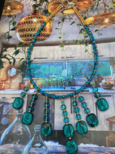 Load image into Gallery viewer, This necklace features vibrant teal quartzite and glass beads. The quartzite beads have a captivating swirl pattern, combining dark red and dark blue hues. It has an ethereal feel thanks to the various shades of blues, golds, and greens used to create a two-strand beaded design. For a dramatic finish, seven strands are added, completed with rectangular rainbow hematite beads for texture and depth. Both strands are held together with double-hole flat matte gold beads to keep a collar shape.