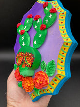 Load image into Gallery viewer, This handcrafted polymer clay wall art features a wood frame painted in a gradient from light to dark purple, creating a stunning sunset look. With a bright green skull cactus plant and bright red blooming flowers, this piece is sure to make a statement. Measurements: H 7" x W 5” x D 1 1/2"