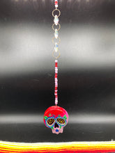 Load image into Gallery viewer, Nature inspired hand crafted polymer clay skull is painted in ombre colors from red, to light pink and decorated with flowers on back and over eyes. Three hearts rainbow hematite bead on forehead. Light pink glass beaded chain with iridescent crystals. A perfect hanging Day of the Dead art piece to add to your ofrenda, hang next to your display of crystals or witchy altar, add to your skull collection or hang as an art piece on the wall or near a window. Measurements: H 1 1/2” x W 1 1/2” x D 3/4”