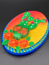 Load image into Gallery viewer, This handcrafted polymer clay wall art features a wood frame painted in a gradient from light yellow to dark red, creating a stunning sunset look. With a bright green skull cactus plant and bright pink blooming flowers. Boasting vivid marigold blooms and a deep royal blue frame to make a bold contrast with the warm background hue. this piece is sure to make a statement. Measurements: H 7" x W 5” x D 1 1/2"