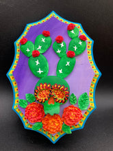 Load image into Gallery viewer, This handcrafted polymer clay wall art features a wood frame painted in a gradient from light to dark purple, creating a stunning sunset look. With a bright green skull cactus plant and bright red blooming flowers, this piece is sure to make a statement. Measurements: H 7" x W 5” x D 1 1/2"