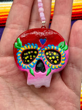 Load image into Gallery viewer, Nature inspired hand crafted polymer clay skull is painted in ombre colors from red, to light pink and decorated with flowers on back and over eyes. Heart rainbow hematite bead on forehead. Light pink glass beaded chain with iridescent crystals. A perfect hanging Day of the Dead art piece to add to your ofrenda, hang next to your display of crystals or witchy altar, add to your skull collection or hang as an art piece on the wall or near a window. Measurements: H 1 1/2” x W 1 1/2” x D 3/4”