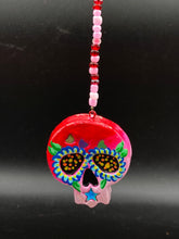 Load image into Gallery viewer, Nature inspired hand crafted polymer clay skull is painted in ombre colors from red, to light pink and decorated with flowers on back and over eyes. Three hearts rainbow hematite bead on forehead. Light pink glass beaded chain with iridescent crystals. A perfect hanging Day of the Dead art piece to add to your ofrenda, hang next to your display of crystals or witchy altar, add to your skull collection or hang as an art piece on the wall or near a window. Measurements: H 1 1/2” x W 1 1/2” x D 3/4”