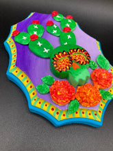 Load image into Gallery viewer, This handcrafted polymer clay wall art features a wood frame painted in a gradient from light to dark purple, creating a stunning sunset look. With a bright green skull cactus plant and bright red blooming flowers, this piece is sure to make a statement. Measurements: H 7" x W 5” x D 1 1/2"