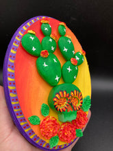 Load image into Gallery viewer, This handcrafted polymer clay wall art features a wood frame painted in a gradient from light yellow to dark red, creating a stunning sunset look. With a bright green skull cactus plant and bright yellow/red blooming flowers. Boasting vivid marigold blooms and a deep purple frame to make a bold contrast with the warm background hue. this piece is sure to make a statement. Measurements: H 7" x W 5” x D 1 1/2"