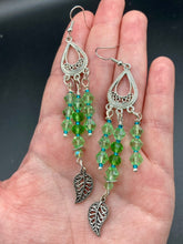 Load image into Gallery viewer, This large set of chandelier earrings features three long strands of shimmering glass beads in various light green and blue hues, accented with an intricate silver-plated leaf charm at the bottom for an added touch of movement. Versatile and perfect for whimsical, hippie-inspired, nature-loving looks. Measurements: 3 3/4” ” in length