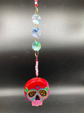 Load image into Gallery viewer, Nature inspired hand crafted polymer clay skull is painted in ombre colors from red, to light pink and decorated with flowers on back and over eyes. Three hearts rainbow hematite bead on forehead. Light pink glass beaded chain with iridescent crystals. A perfect hanging Day of the Dead art piece to add to your ofrenda, hang next to your display of crystals or witchy altar, add to your skull collection or hang as an art piece on the wall or near a window. Measurements: H 1 1/2” x W 1 1/2” x D 3/4”